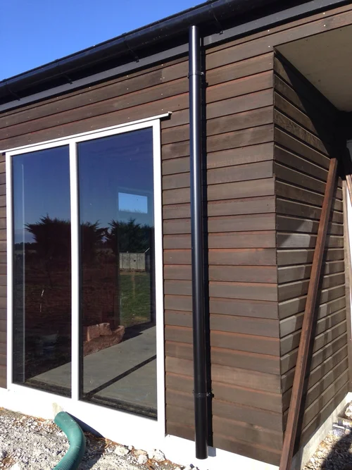 Exterior view of a modern building with dark wood siding, large windows, and a black downspout. A metal beam leans against the wall.