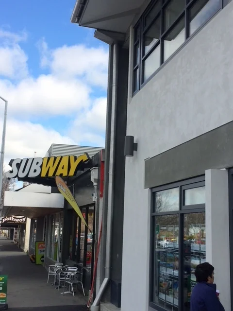 Subway restaurant storefront on a sidewalk, next to a building with windows. Blue sky with clouds overhead.