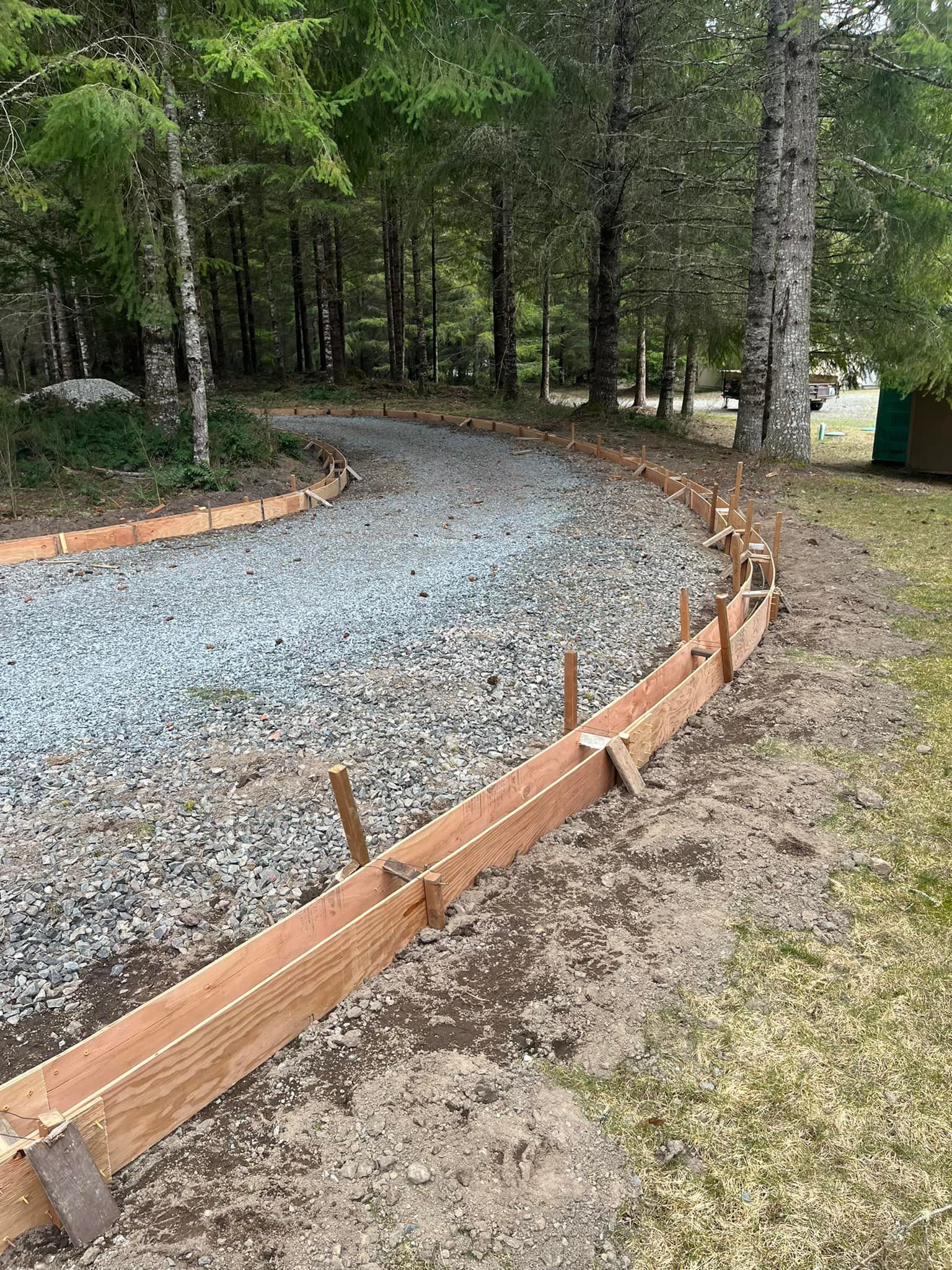 Gravel driveway with wooden forms for concrete edging, bordered by grass and trees.