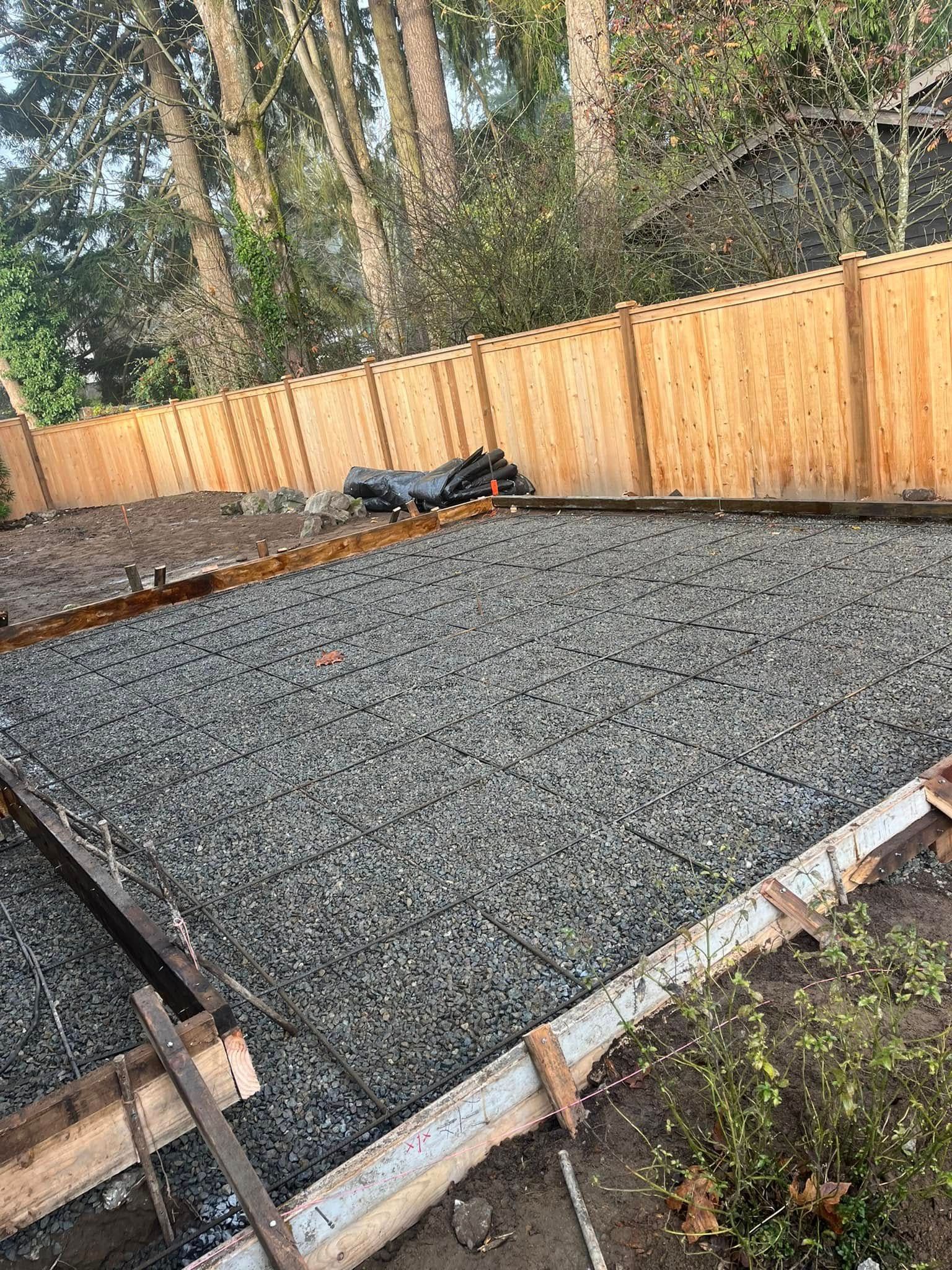 Gravel filled form for concrete base in backyard, surrounded by wooden fence and trees.