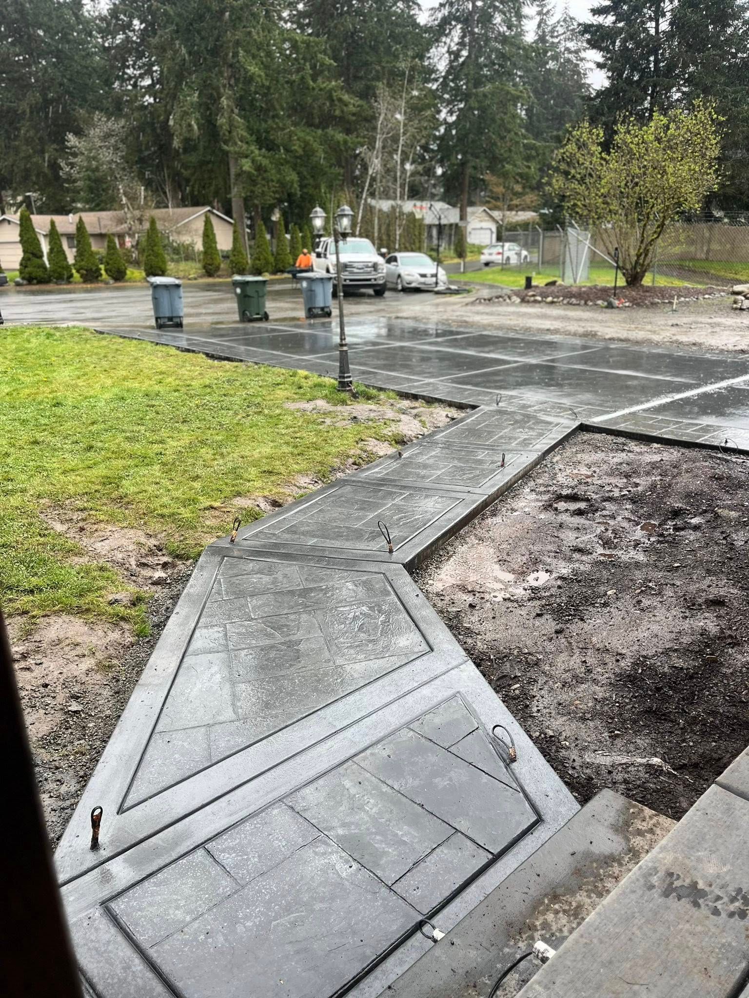 Exterior view of a paved area with a walkway leading to a parking lot, with green grass on the left and trees in the background.
