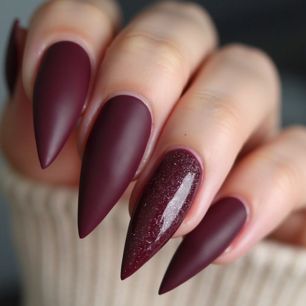 Close-up of hand with long, maroon matte stiletto nails, one with glitter.