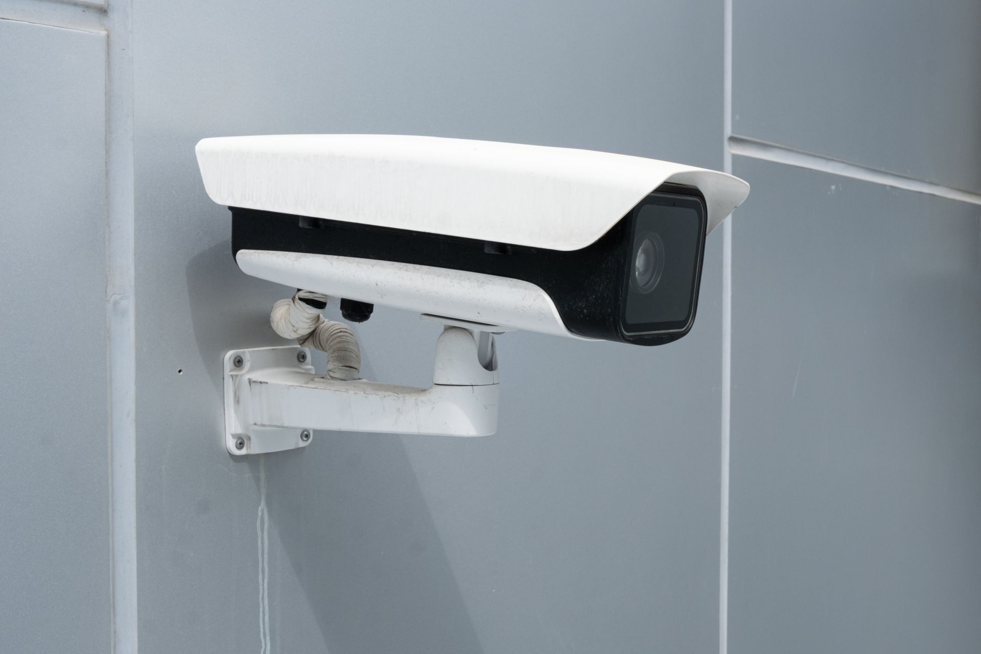 White security camera mounted on a gray wall, angled downward outdoors.