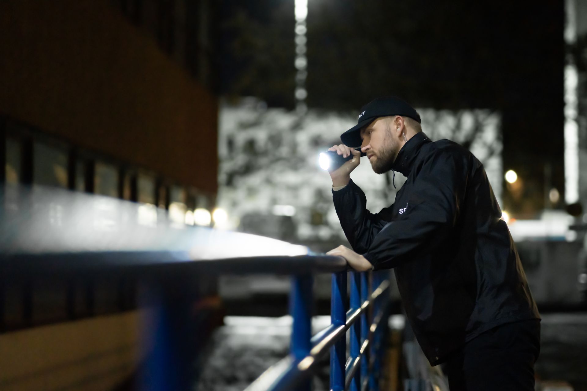 Man in black jacket and cap leaning on a railing, looking into a phone at night.
