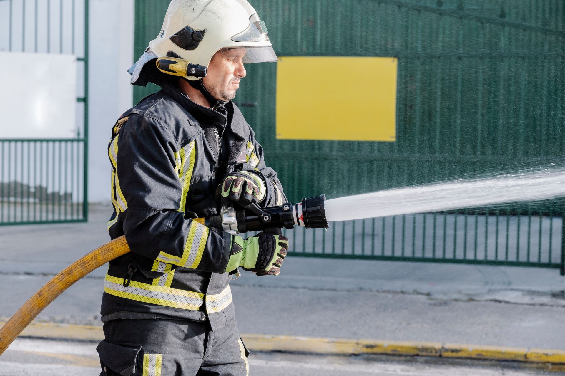 Firefighter in black gear spraying water from a hose outside a building.