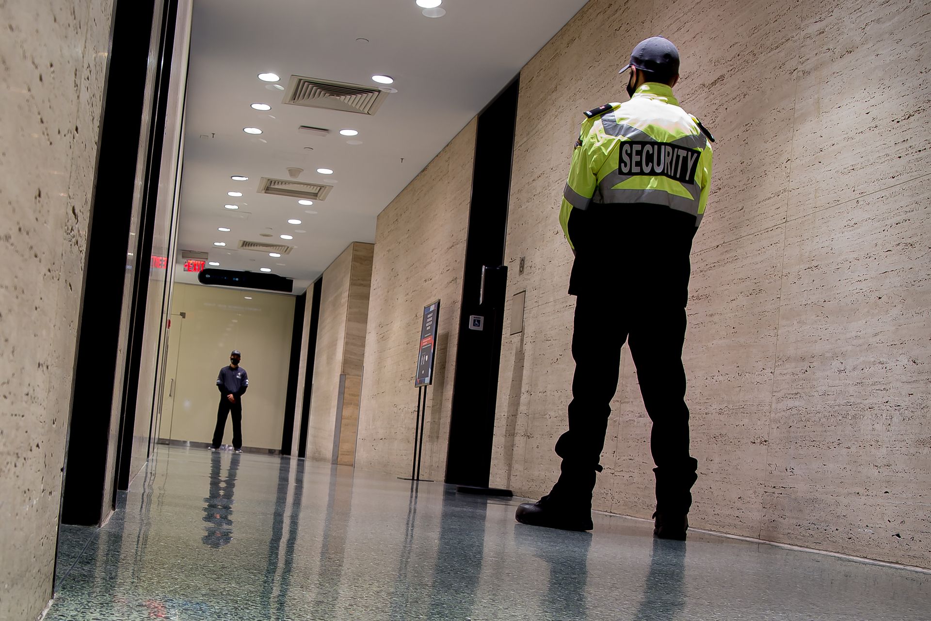 Security guard standing in a hallway, facing a distant person under bright ceiling lights.