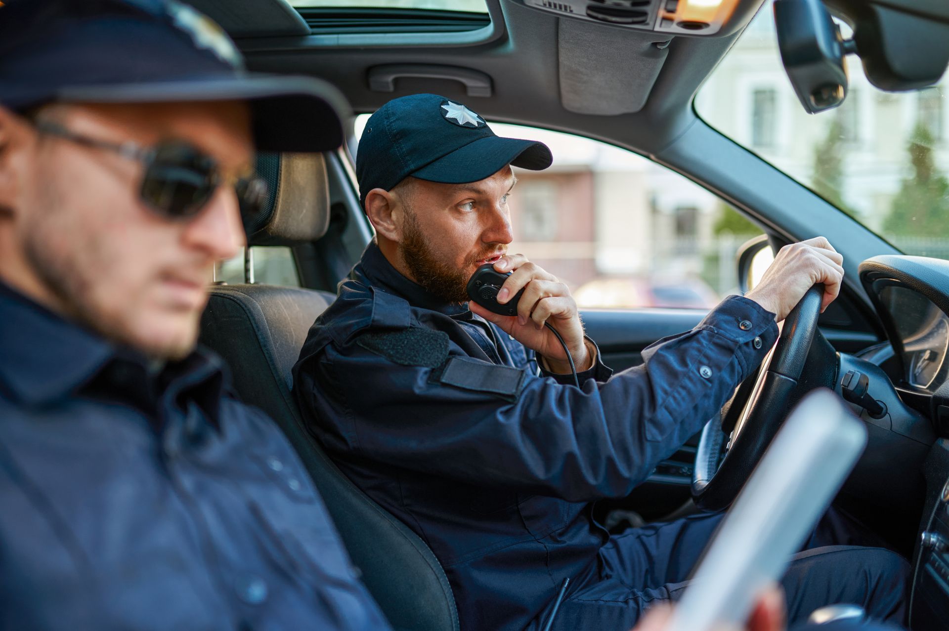 Two police officers in uniform inside a patrol car, one driving and one reading a device.