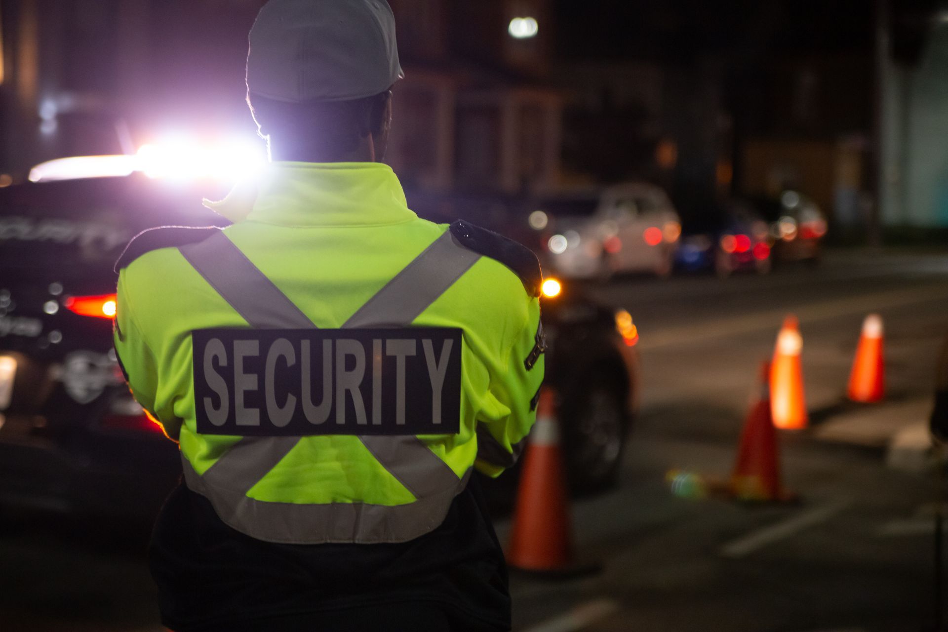 Security guard in a neon vest standing near traffic cones at night with flashing lights nearby.