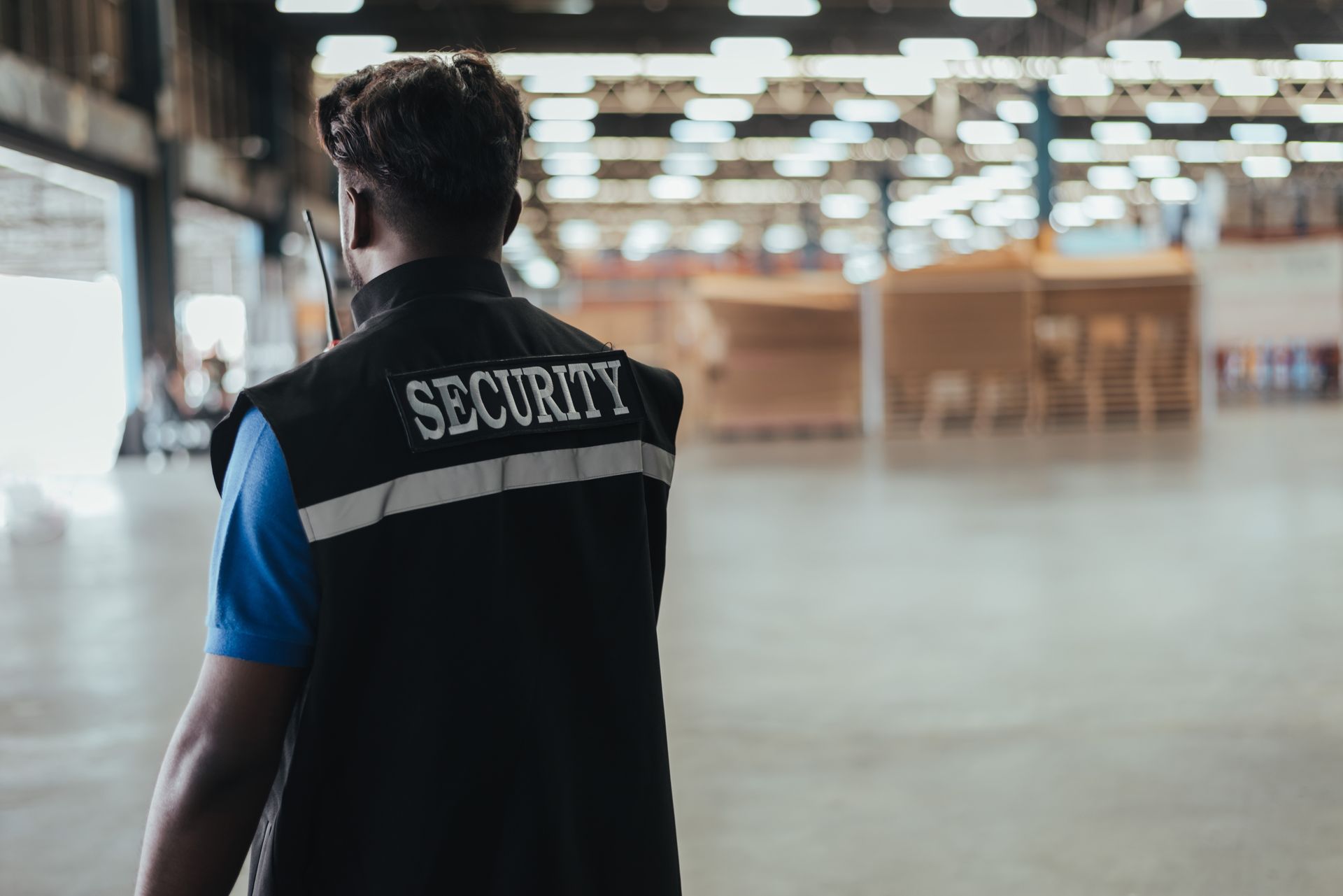 Security guard walking through an empty indoor hall, wearing a black vest with “SECURITY” on the back.