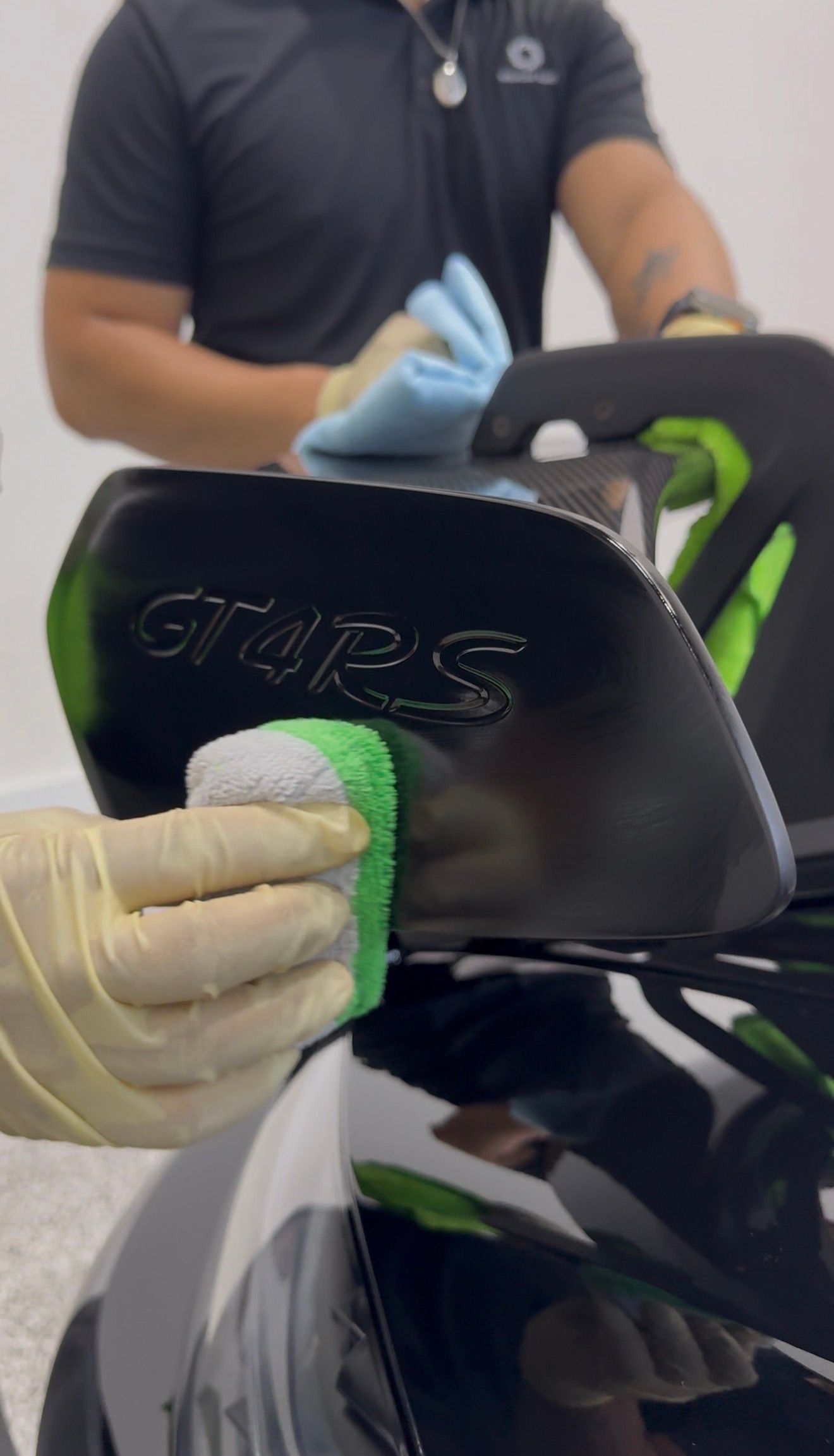 A person wearing gloves uses an applicator pad to apply a coating to the GT4 RS wing of a black car.