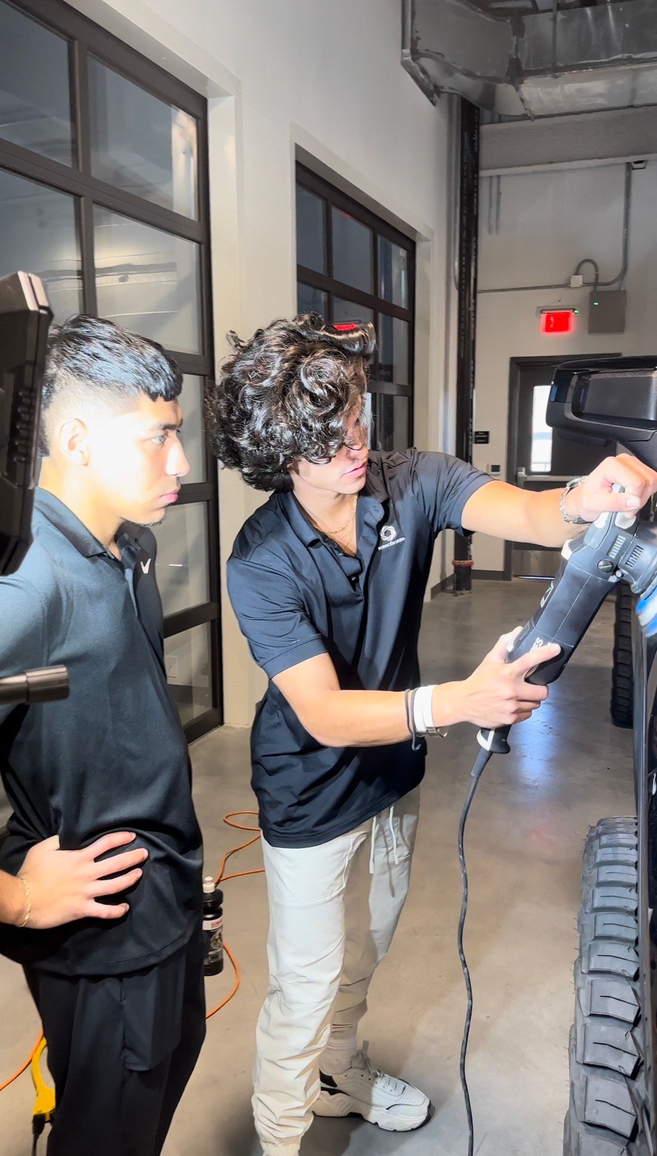 Two men examine equipment in a workshop. One points; the other observes. The setting has large windows.