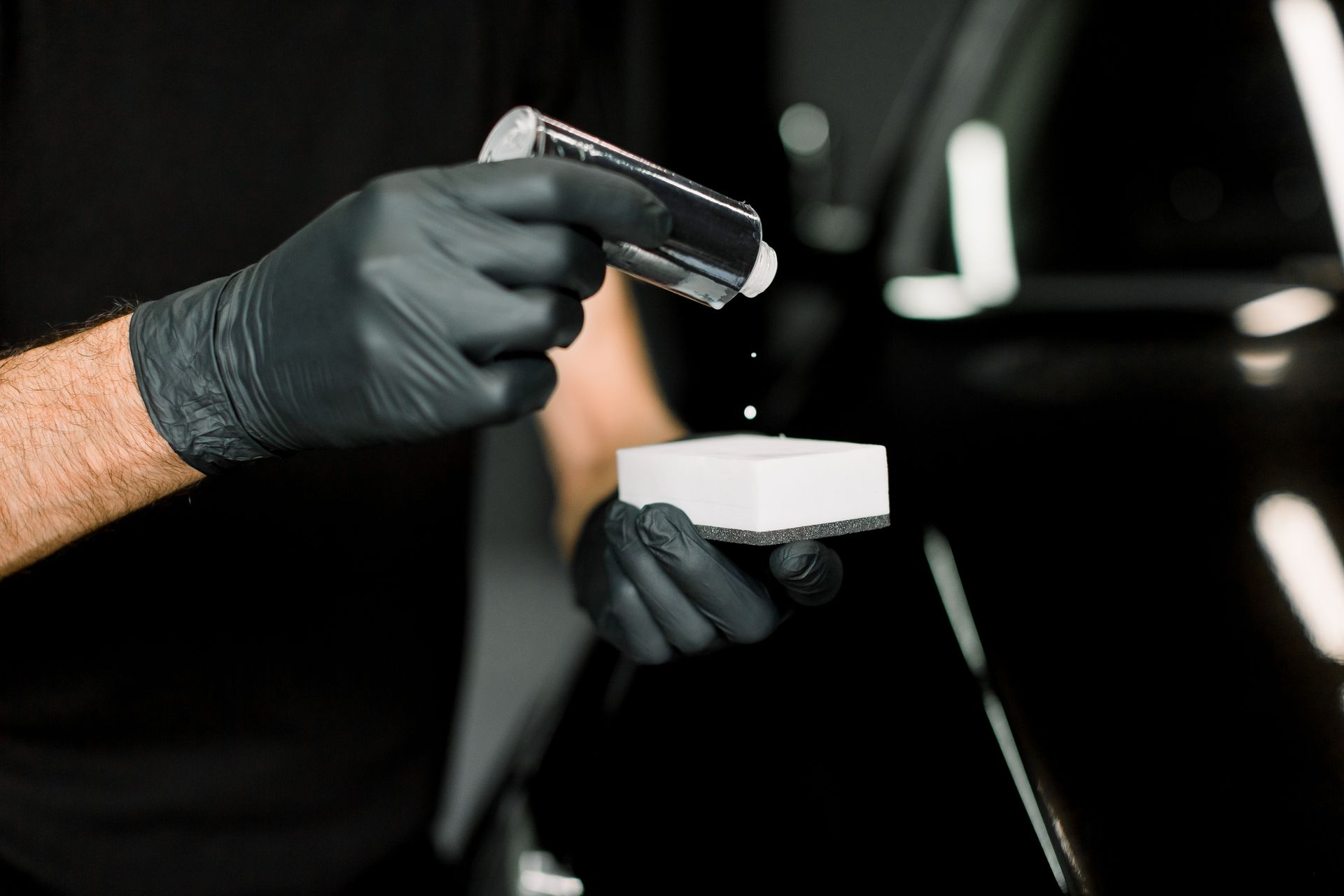 Gloved hands holding a spray bottle and sponge, cleaning a black car door.