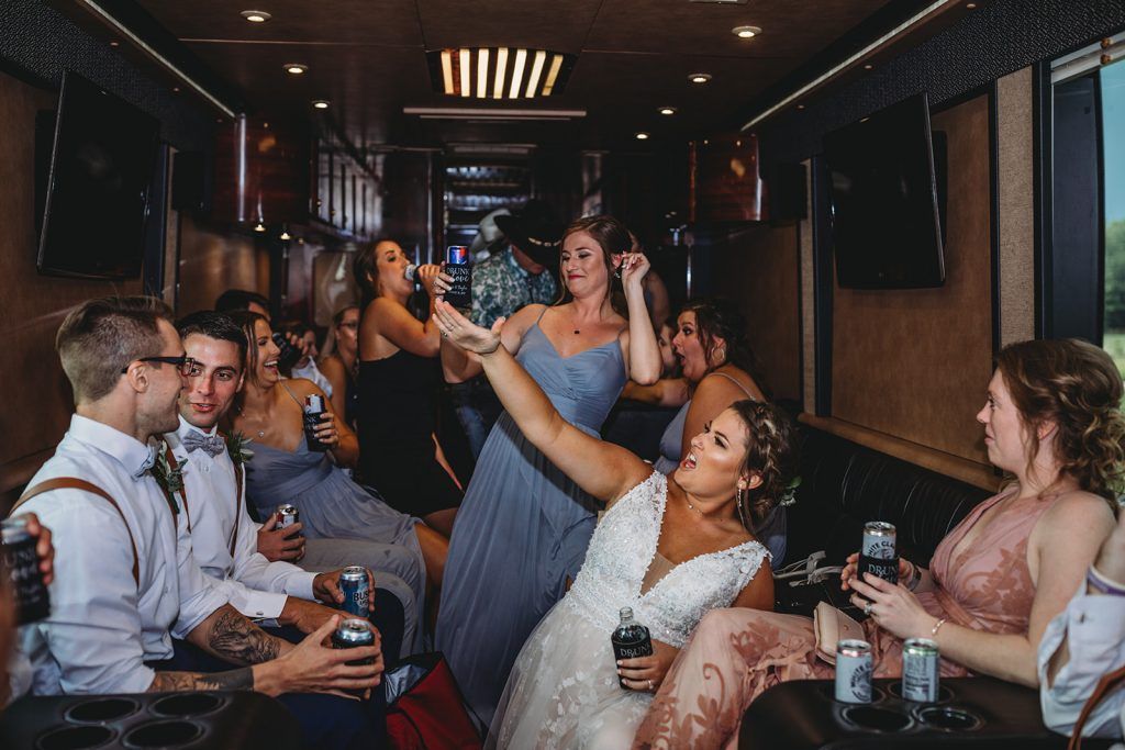 People celebrating inside party bus, some drinking. Bride in white dress dancing, others smiling. Dark interior with screens