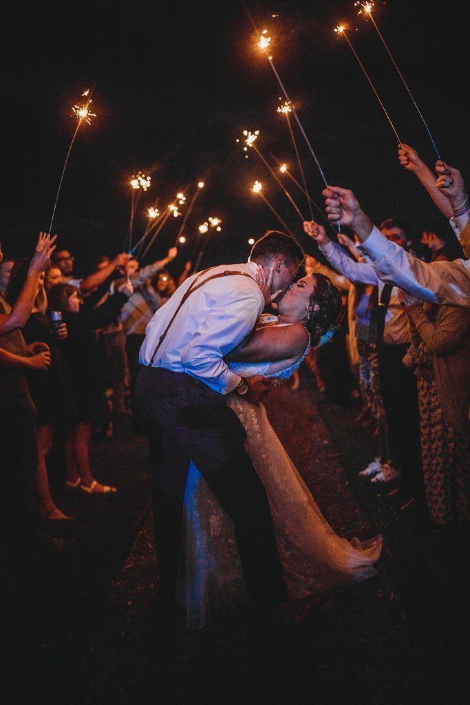 Newlyweds kiss under a sparkler tunnel at night. Guests hold lit sparklers, celebrating.
