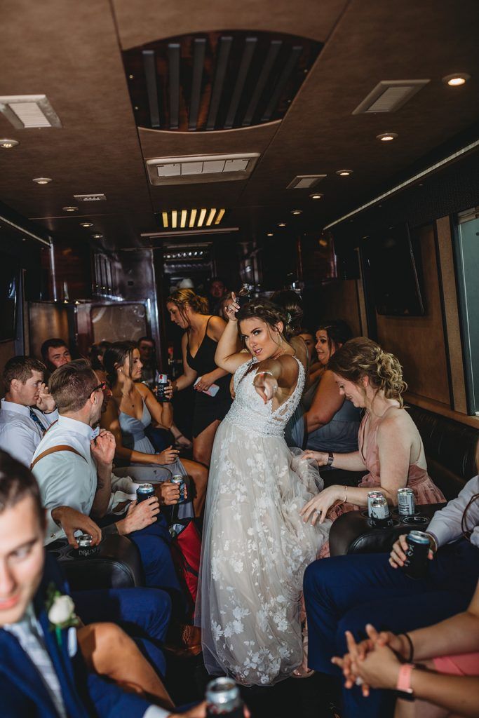 Bride dances in a party bus with wedding guests, drinks in hand. Brown interior, lots of smiling faces.