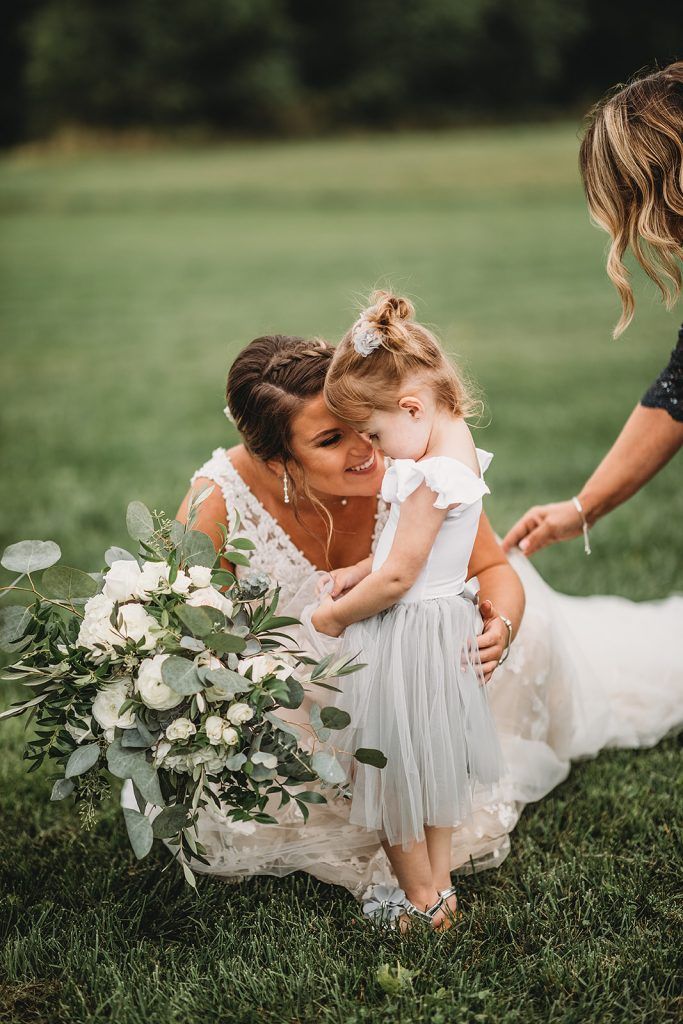 Bride kneeling, hugging flower girl, holding bouquet in grassy field; woman's hand on girl's back.