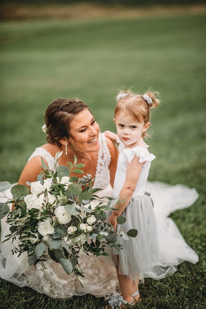 Bride with a bouquet smiles at a child flower girl in a grassy field.