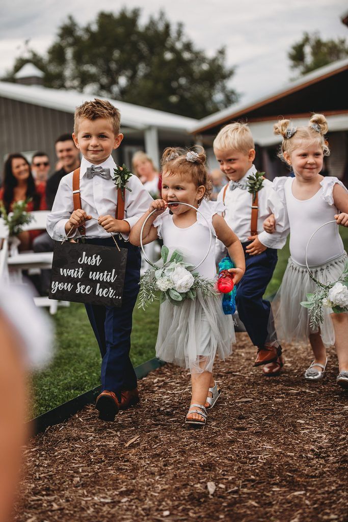 Children in wedding attire walking down a path; one holds a sign, others flowers.