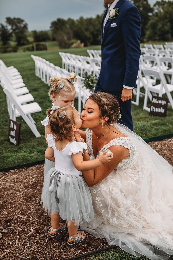 Bride kissing two flower girls at a wedding ceremony. White chairs line the grass.