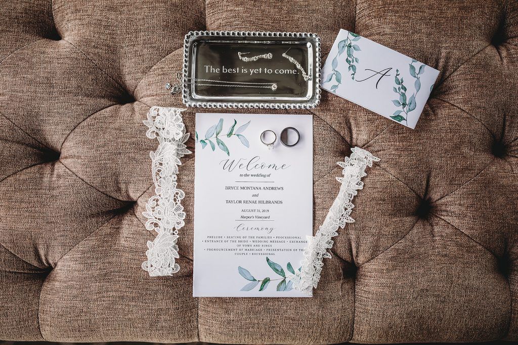 Wedding invitation suite with rings, lace, and silver tray on tufted surface.
