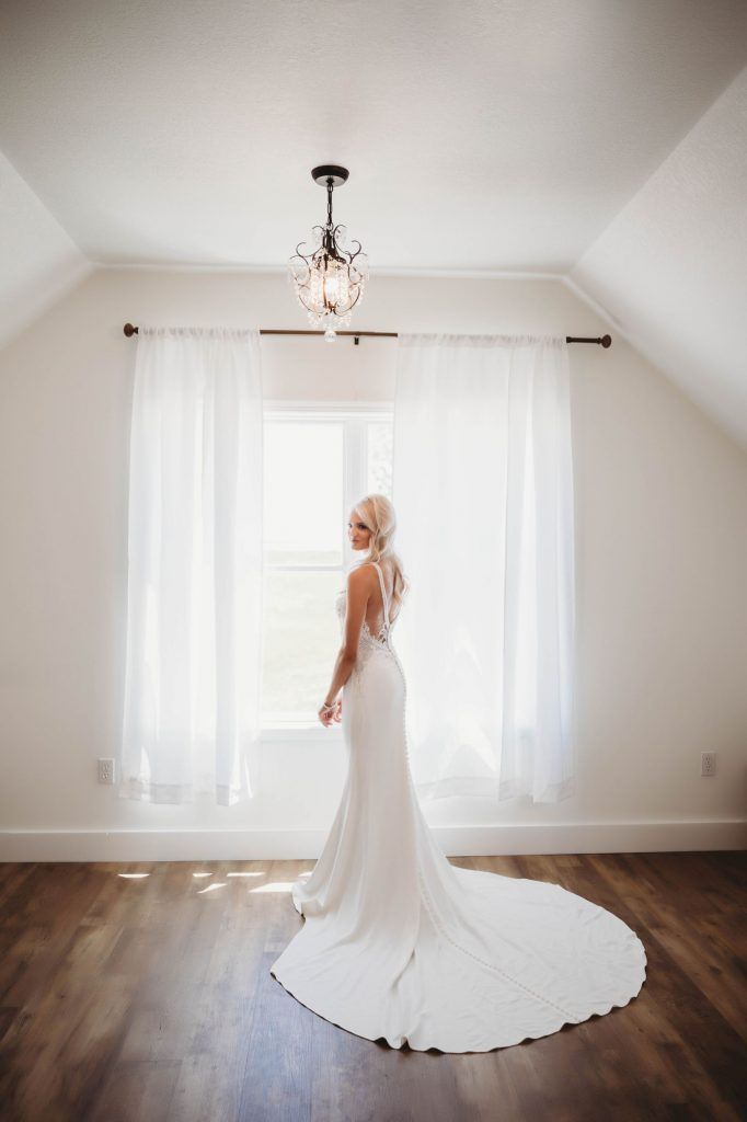 Bride in a white wedding dress stands near a window in a bright room, looking over her shoulder.