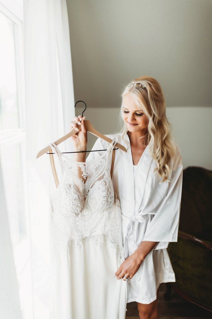 Woman in white robe holding up a wedding dress, smiling, next to a window.