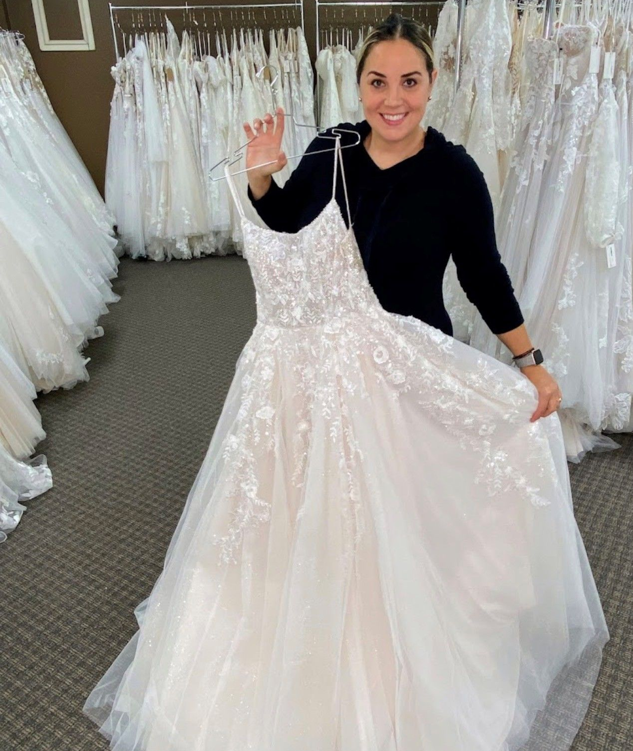 Woman in a bridal shop holding a wedding dress, smiling and waving.