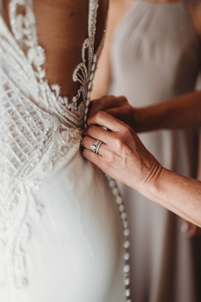 A person's hands buttoning a white wedding dress. Another person stands beside.
