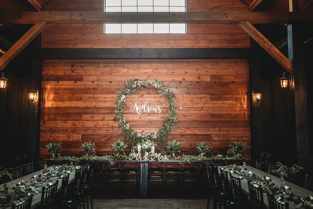 Wedding reception: Long tables with greenery in a wooden barn, garland and floral backdrop, overhead lighting.