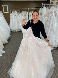 Woman in a bridal shop holding a wedding dress, smiling and waving.