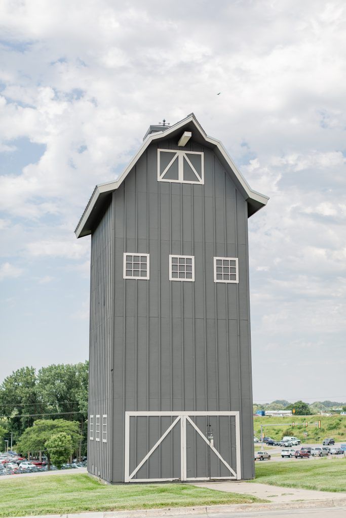 Tall, gray barn with white trim, three windows, and double doors. Green grass and parked cars in the background.