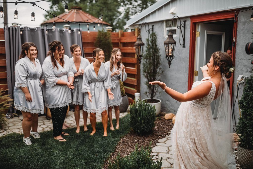 Bride throwing confetti; bridesmaids watch. Garden setting with string lights and small building.