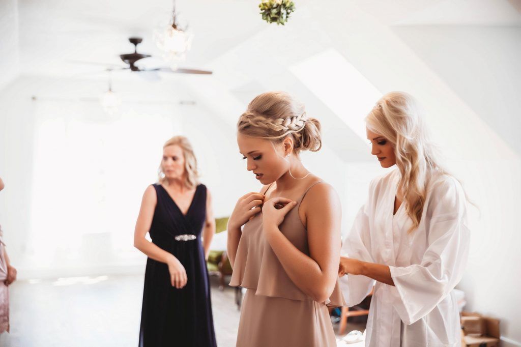 Bride getting help with dress by woman in robe, other woman watches. Bright room.