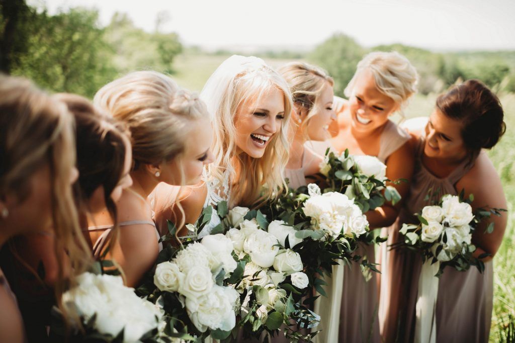 Bride laughs with bridesmaids in a field, holding white bouquets, wearing tan gowns.