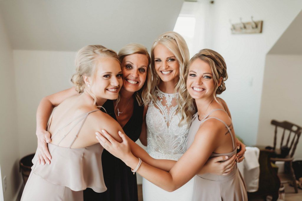 Bride surrounded by bridesmaids hugging in a room, smiling. Soft beige and black dresses, natural light.