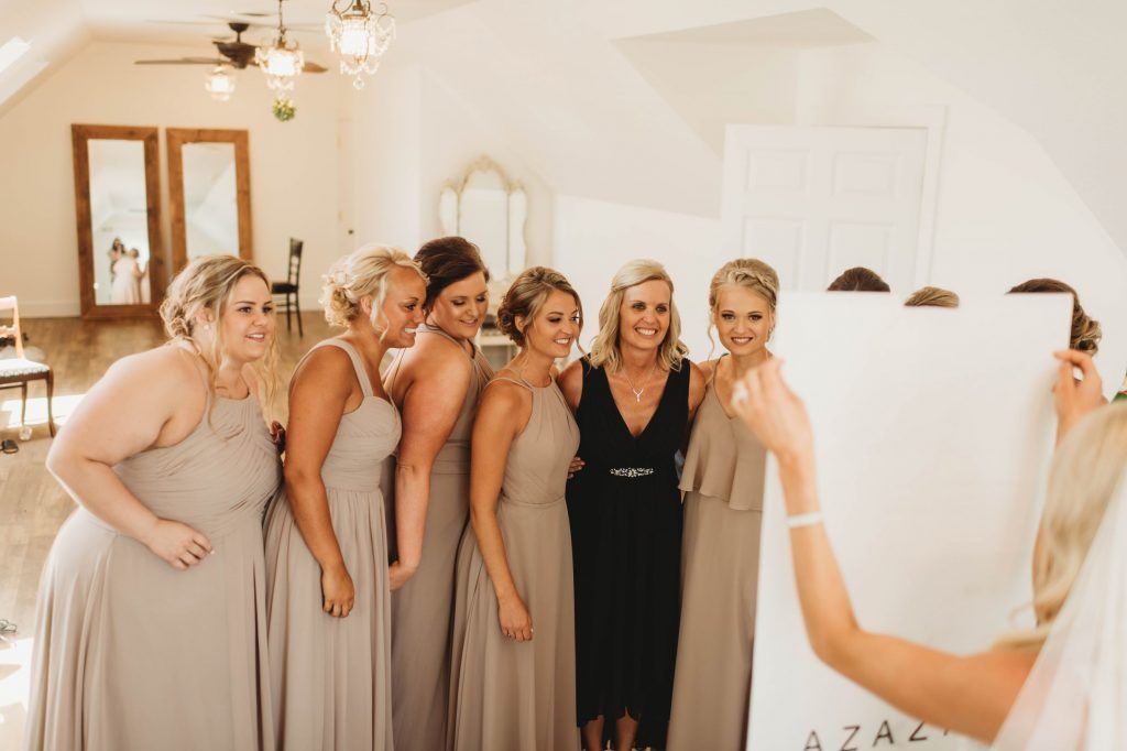 Bridesmaids in taupe dresses pose with a woman in black. One holds a sign while another takes a photo in a bright room.