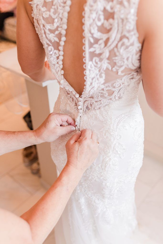 Woman's back in a white lace wedding dress being buttoned up by someone's hands.