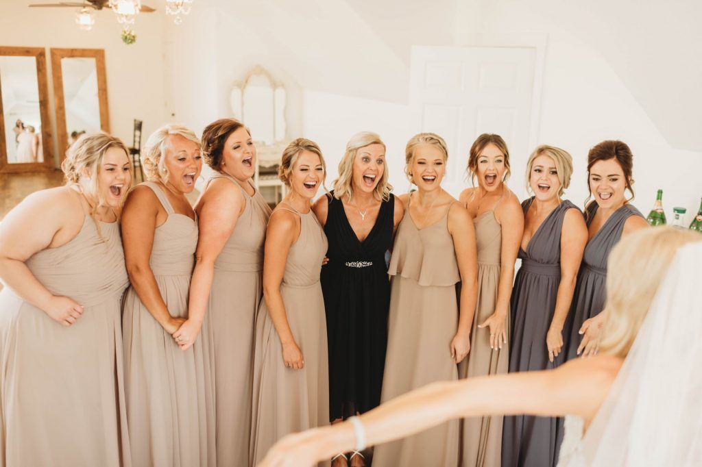 Bridesmaids cheering at bride wearing a veil, indoors. Beige, gray dresses, light room.