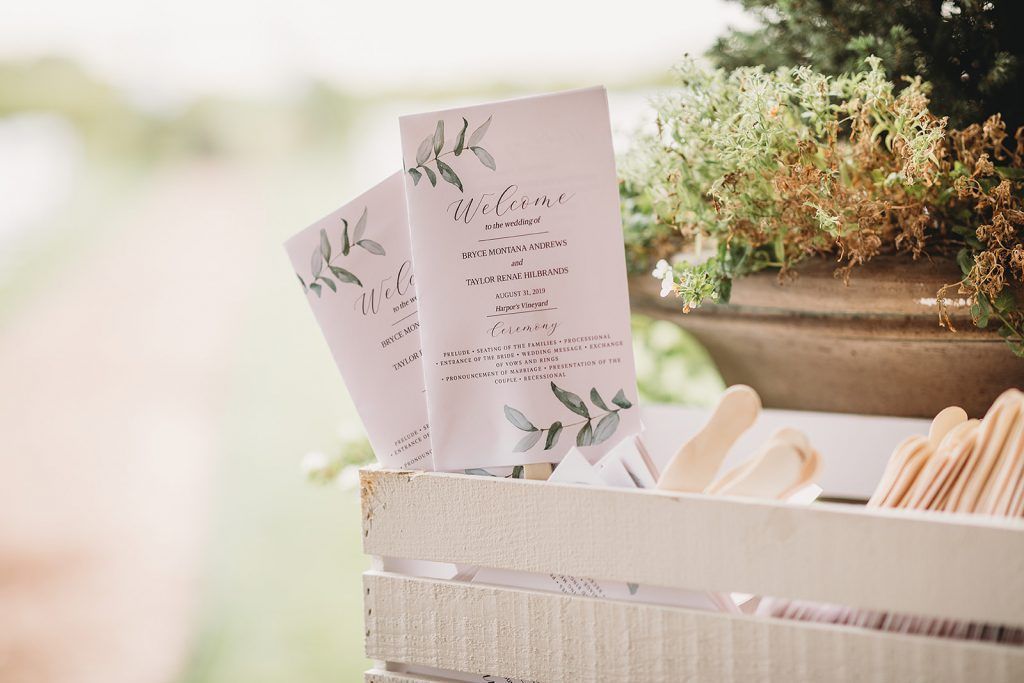 Two wedding programs with green leaf design in a white wooden crate next to wooden fans.