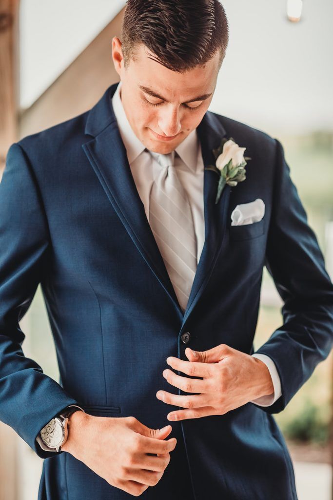 Man in navy blue suit buttoning his jacket, white tie, pocket square, and watch.