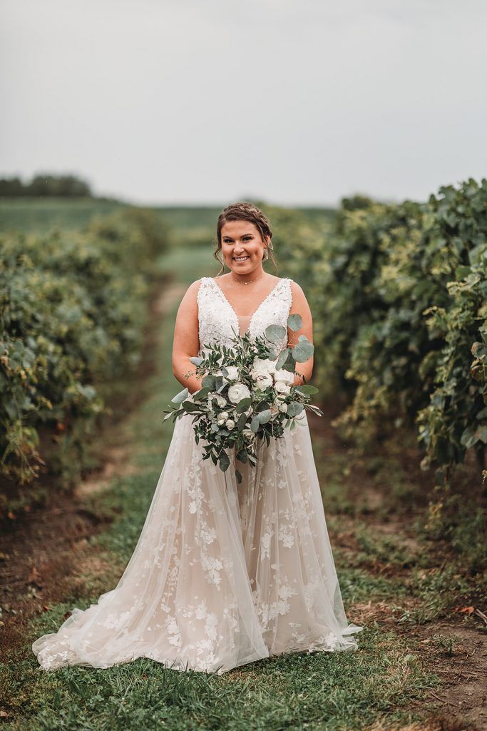 Bride in white lace dress holding bouquet, standing in a vineyard.