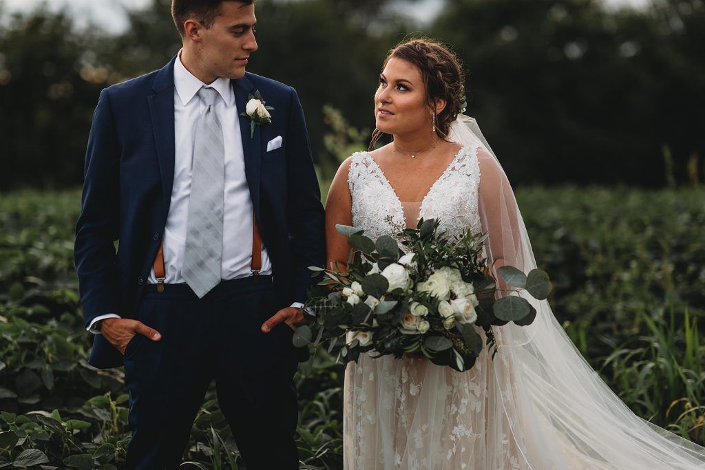Groom in navy suit and bride in white dress, looking at each other in a field.