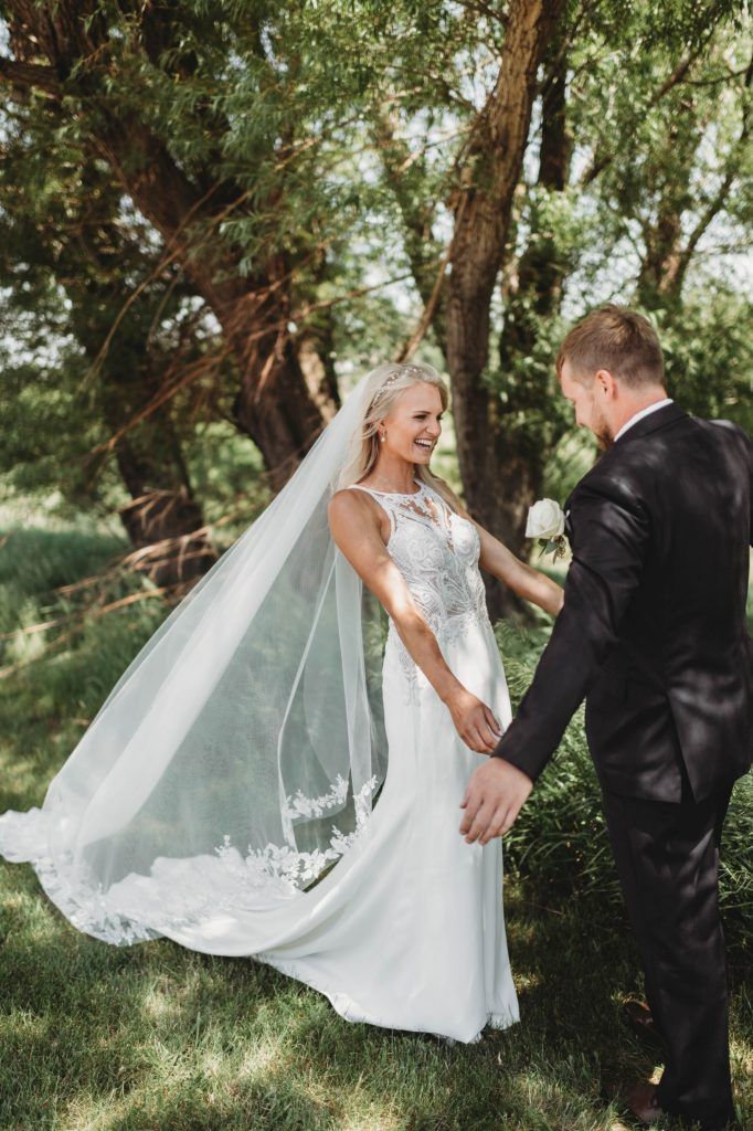 Bride and groom holding hands, smiling. Wedding dress with long veil, outdoors near trees.