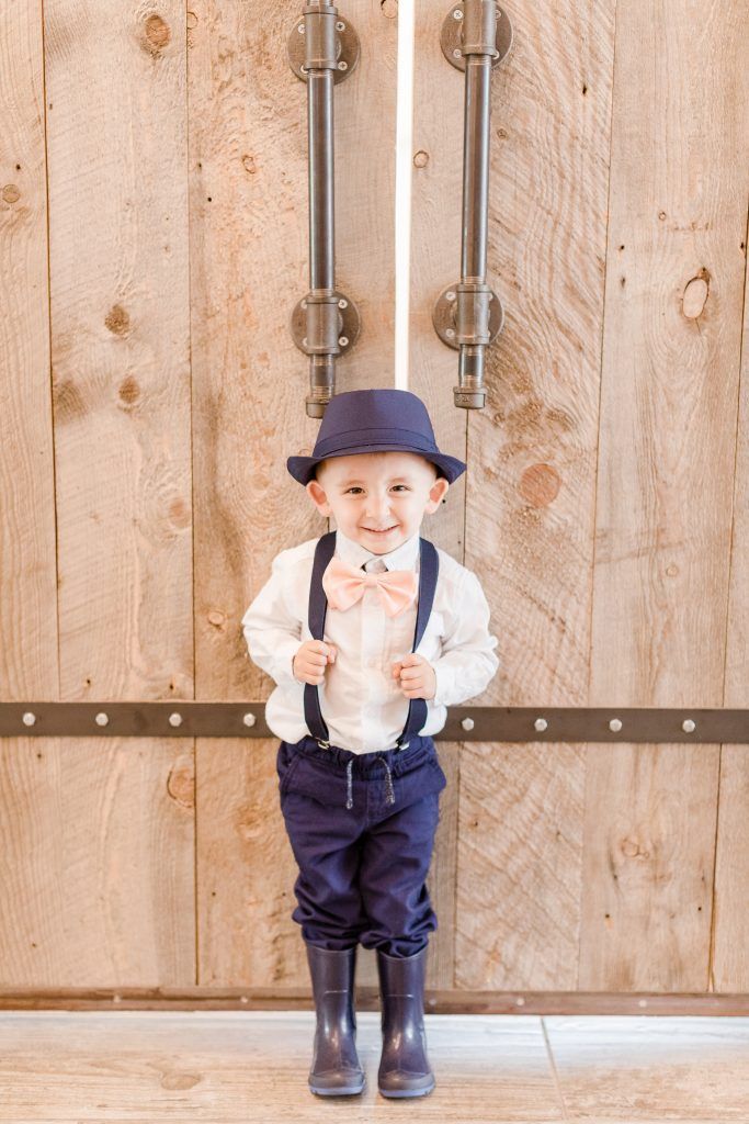 Young child in a hat, suspenders, and bow tie smiles in front of a wooden door with metal hardware.