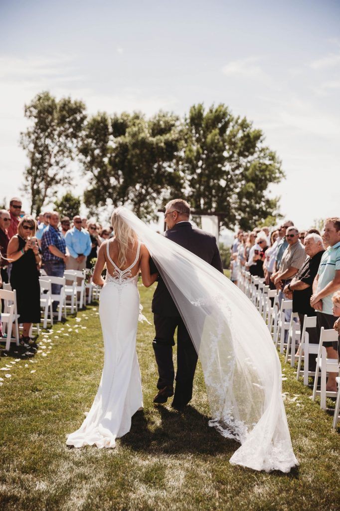 Bride in white dress walks down aisle with father, veil trailing. Wedding ceremony outdoors.