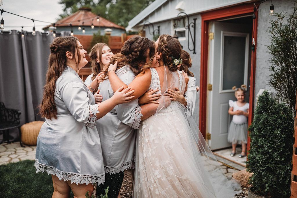 Bride hugs bridesmaids outside a building. They are in a garden setting. A child watches from the doorway.