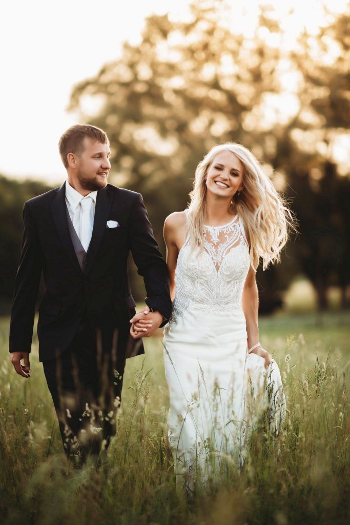 Bride and groom holding hands, smiling, walking through a field of tall grass. Golden light.