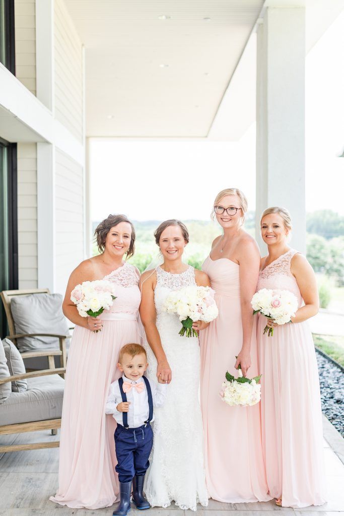Bride and bridesmaids in pink dresses with a boy, posing on a porch, holding bouquets.