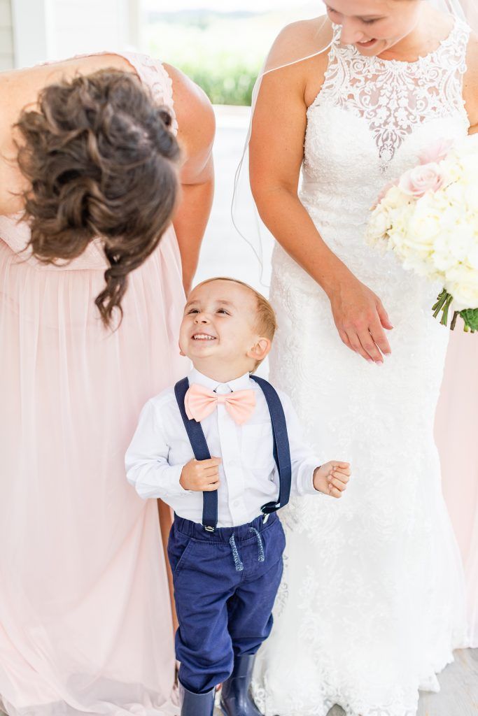 Young boy in bow tie and suspenders smiles, bride and bridesmaid in background.
