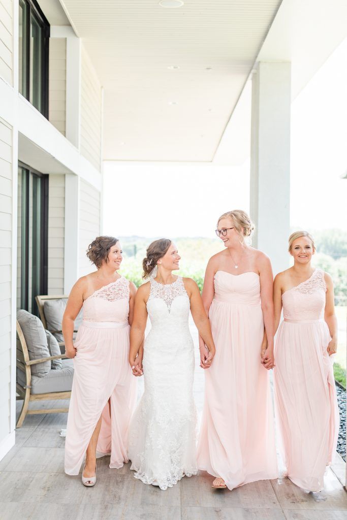 Bride in white dress holds hands with bridesmaids in pink dresses on a porch.